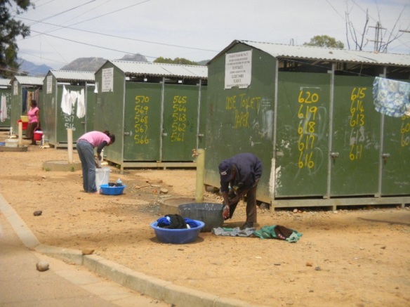 South Africa ghetto restrooms.  Each outhouse has 5 house numbers painted on it to show which 5 families share the toilet.  Women wash clothes at the water spigots.